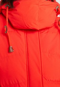 Czerwona pikowana kurtka puchowa z wysokim kołnierzem, grube przeszycia, sznurki oraz brązowe akcenty z troczkami do regulacji przy szyi.