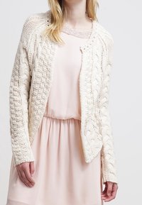 Vit stickad cardigan med kabelmönster, öppen fram, och långa ärmar, bärs över en mjuk rosa klänning med en strukturerad halsringning.