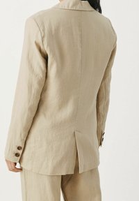 Beige pellavapuvun takki, jossa on hieman teksturoitu pinta, lovikaulus ja takakara, sekä nappikoristeet hihoissa.