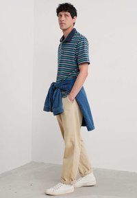 Cămașă polo în dungi, de culoare navy, teal și albastru deschis; asortată cu pantaloni bej și o jachetă din denim legată în jurul taliei. Adidași albi.