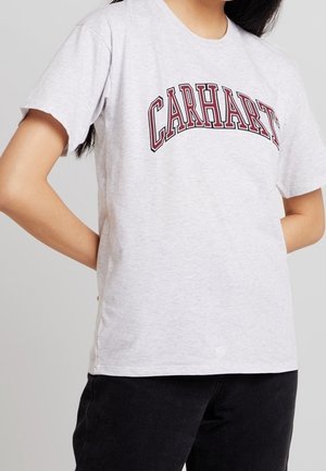 Person iført en lysegrå Carhartt T-shirt med rødt omridset tekst og sorte bukser, stående med hænderne bag ryggen.