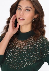Zielony sweter z dzianiny o wysokim kołnierzu, ozdobiony rozrzuconymi złotymi cekinami z przodu. Modelka prezentuje neutralny makijaż.