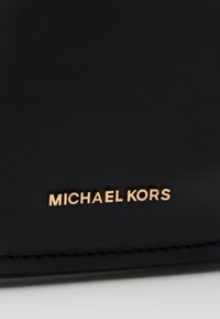 Svart lädermaterial med en jämn textur, prytt med en guld "MICHAEL KORS"-logotyp som finns i framkant. Klassisk design och minimalistisk detaljering.
