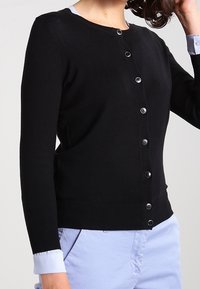 Svart cardigan med rund halsringning, långa ärmar och knappstängning framtill. Har ribbade muddar och fåll, tillverkad av mjukt stickat material.