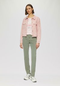 Rosa Tweed-Jacke mit Fronttaschen und großen Knöpfen, kombiniert mit einem hellrosa Grafik-T-Shirt und hellgrünen Hosen, abgerundet mit grauen Turnschuhen.