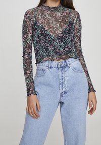 Top nero trasparente a maniche lunghe con motivo floreale in rosa e verde, rifinito con dettagli smerlati, abbinato a jeans azzurri a vita alta.