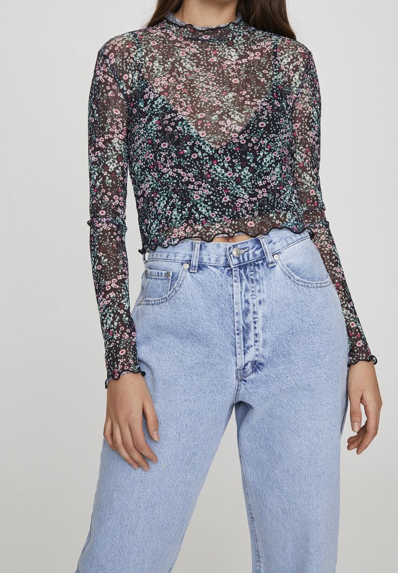 Top nero trasparente a maniche lunghe con motivo floreale in rosa e verde, rifinito con dettagli smerlati, abbinato a jeans azzurri a vita alta.