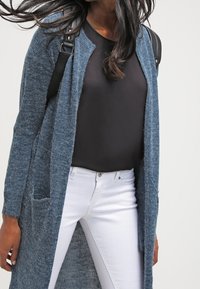 Cardigan lungo aperto di maglia strutturata in blu navy, con due tasche anteriori, indossato sopra un top aderente nero e jeans bianchi.