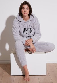 Sivi krzneni kombinezon s kapuljačom, s printom "MEOW" i ušima mačke. Mekana tekstura, duge rukave i sužene nogavice.