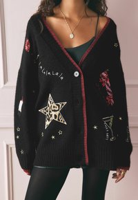 Cárdigan negro con ribetes rojos, que presenta una estrella de estampado de leopardo y detalles bordados coloridos, incluyendo bebidas y estrellas. Cuatro botones.