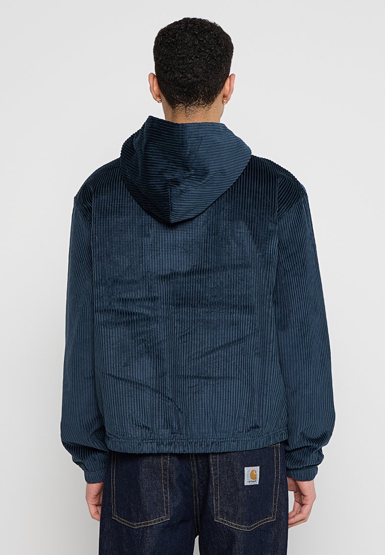 Veste à capuche en velours côtelé marine avec un tissu texturé, un motif à côtes et des poignets élastiques ; portée par-dessus un pantalon en jean foncé avec des coutures visibles.
