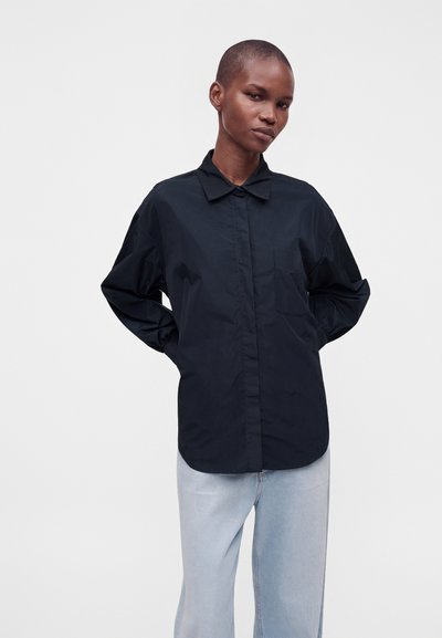Personne aux cheveux courts portant une chemise noire ample à boutons et un jean bleu clair, debout les mains derrière le dos sur fond blanc.
