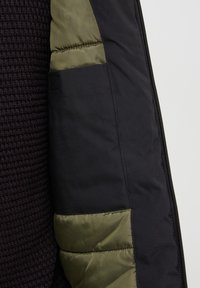 Giacca nera con texture lavorata a maglia, manica nera liscia e dettagli verde oliva nella fodera, dotata di zip.