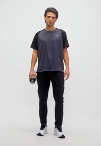 Νέος άνδρας με αθλητική ενδυμασία Puma σε μαύρο και γκρι, κρατώντας ένα διαφανές παγούρι με νερό, στέκεται πάνω σε ανοιχτό γκρι φόντο.