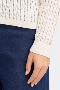 Pull en crème tricoté avec un motif ajouré et des poignets côtelés, associé à un denim bleu foncé. Accent sur la texture et les détails de design.
