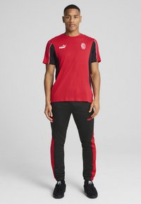 Červené športové tričko s čiernymi rukávmi, s logom Puma; čierne nohavice s červenými bočnými panelmi a logom Puma vpredu.