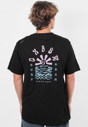 Personne aux cheveux bouclés portant un T-shirt noir avec un motif abstrait de soleil et de vagues en pastel rose et bleu avec le texte "Surf Gear Since 1985."