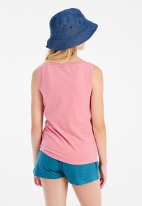 Top senza maniche rosa abbinato a pantaloni corti texturizzati turchesi; un cappello a secchiello blu completa l'outfit, visto da dietro.