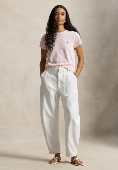 Γυναίκα με μακριά σγουρά μαλλιά, που φοράει ροζ ανοιχτόχρωμο μπλουζάκι, λευκό ψηλόμεσο παντελόνι και λευκά σανδάλια, στέκεται με τα χέρια στις τσέπες.