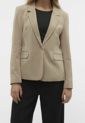 Kobieta ubrana w beżową marynarkę na jeden guzik, na czarnym topie i czarnych spodniach, stojąca z rękami swobodnie wzdłuż ciała.