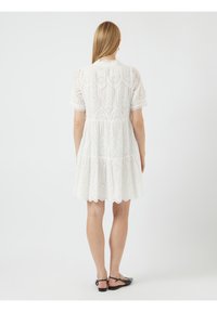 Vit bomullsklänning med korta puffärmar, en volangkant och dekorativ öljettmönster. Har en hög krage och utsvängd silhuett.