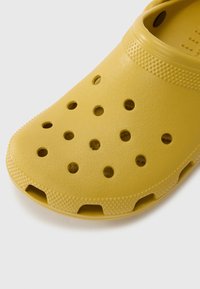 Yellow plastic clog with perforations, textured surface, open heel, rounded toe, and raised dots for grip. Designed for ventilation and comfort.
