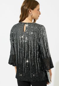 Top nero in paillettes con una superficie testurizzata e scintillante. Presenta maniche a tre quarti e un dettaglio a forma di chiave nella parte posteriore. Abbinato a una pochette argentea.