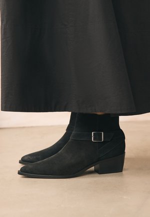 Schwarze Wildleder-Stiefeletten mit niedrigen Blockabsätzen und silbernen Schnallenriemen, getragen mit einem langen schwarzen Rock auf einem beigen Boden.