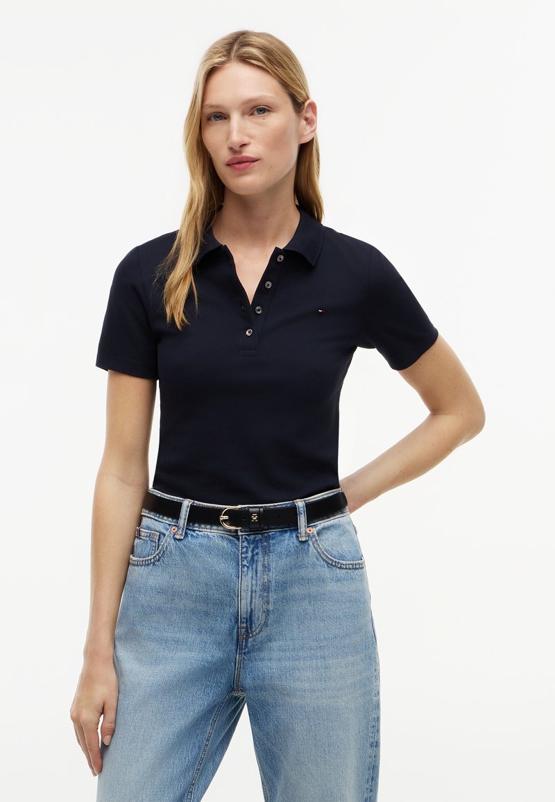 Kvinde med langt blondt hår iført en sort kortærmet polo shirt og lyseblå jeans med sort bælte, står med den ene hånd på hoften.