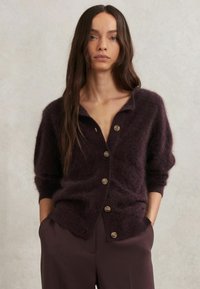Tmavo fialový kardigán vyrobený z mäkkého, nadýchaného materiálu s predným zapínaním na gombíky, golierom a voľným strihom. Obsahuje veľké, okrúhle gombíky.