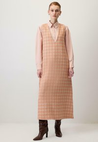 Orange-checkered sleeveless dress with a V-neck, worn over a light pink blouse, paired with brown knee-high heeled boots. Smooth fabric texture.