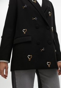 Blazer nero doppiopetto con finitura testurizzata, impreziosito da motivi a cuore e di libellula in oro e perle. Include tasche laterali.