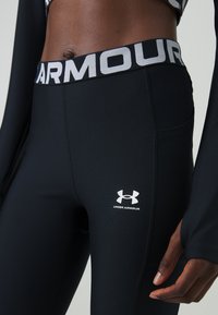 Črne legice z širokim pasom, ki ima napis "ARMOUR" v beli barvi. Logotip Under Armour je viden na spodnjem delu noge. Gladka tekstura.