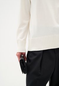 Maglione di crema con orlo e polsini a costine, sovrapposto a pantaloni neri. Una persona tiene una pochette nera, che evidenzia una texture liscia e un design elegante.