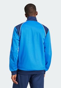 Mies, jolla on sininen ja tummansininen urheilutakki ja housut, seisoo selin kameraan yhtenäisen vaalean taustan edessä.