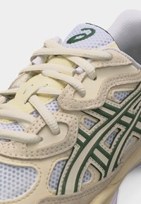 Primer plano de una zapatilla beige y blanca con el logo verde de Asics, tejido de malla, superposiciones de ante y cordones atados sobre un fondo liso.