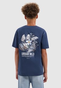 Persoon met krullend haar die een marineblauw T-shirt draagt met een afbeelding van een witte bloem die door beton breekt, met de tekst "Urban Wild."