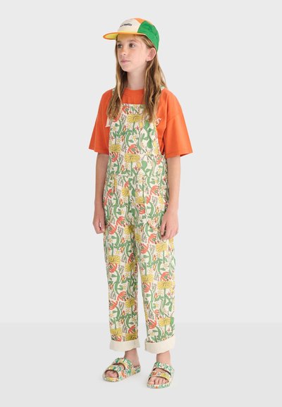 Jeune personne portant une combinaison florale verte, orange et jaune avec des sandales assorties, un t-shirt orange et une casquette multicolore, se tenant face à l'avant.