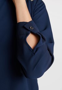 Námořnickomodrá košile s ohrnutým rukávem a detaily s kovovým drukem. Hladká tkanina s jemným leskem a volný střih.