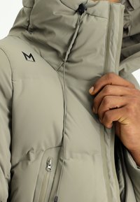Padded jacket w jasnym oliwkowym kolorze, z wysokim kołnierzem, regulowanym kapturem, zamykana boczną kieszenią i czarnym logo na klatce piersiowej.