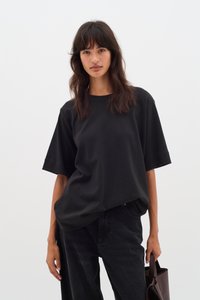 T-shirt nera oversize in tessuto leggero, con maniche corte e scollo rotondo, abbinata a pantaloni in denim scuro e una borsa a tracolla marrone.