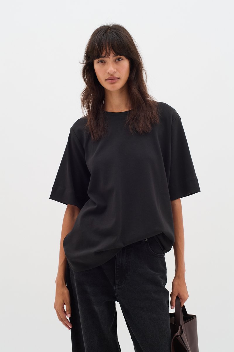 T-shirt nera oversize in tessuto leggero, con maniche corte e scollo rotondo, abbinata a pantaloni in denim scuro e una borsa a tracolla marrone.