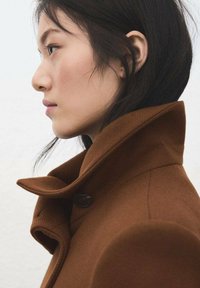 Bruine wollen overjas met een hoge kraag, voorzien van een grote knoop en gestructureerde stof. Zijaanzicht met nadruk op de vorm en details van de kraag.