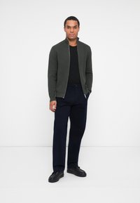 Pull zip vert avec un col haut, porté par-dessus une chemise noire. Assorti à un pantalon bleu marine foncé et des chaussures noires. Texture lisse, design simple.