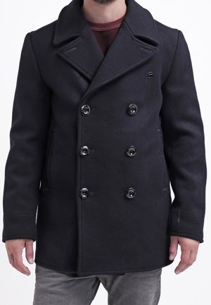 Mann trägt einen schwarzen doppelt geknöpften Peacoat mit sechs Knöpfen, darüber ein dunkelrotes Hemd und graue Hose, steht vor weißem Hintergrund.