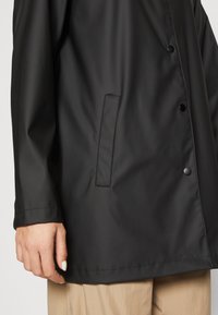 Czarna wodoodporna kurtka z zapięciem na zatrzaski, gładką fakturą i boczną kieszenią. Model charakteryzuje się luźnym krojem i długimi rękawami.
