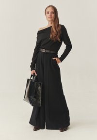 Juodas off-shoulder viršus derinamas su laisvomis juodomis kelnėmis, susiaurintomis rudos spalvos diržu. Modelis laiko juodą odinę rankinę ir ant kojų turi smailius rudus batus.