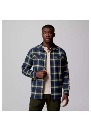 Plaid flannel shirt with green, navy, and cream colors, featuring buttoned pockets and a collar, worn over a beige shirt.