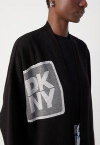 Crna kardigan s otvorenim prednjim dijelom i sivim DKNY logom s lijeve strane. Mekana tekstura i opušteni kroj, nosi se preko crne majice s plavim trapericama.