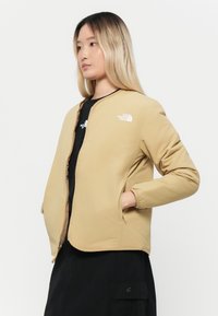 Veste zippée beige avec un col rond et des poignets élastiques. Présente un logo sur la poitrine, des poches latérales et une texture lisse et légère.
